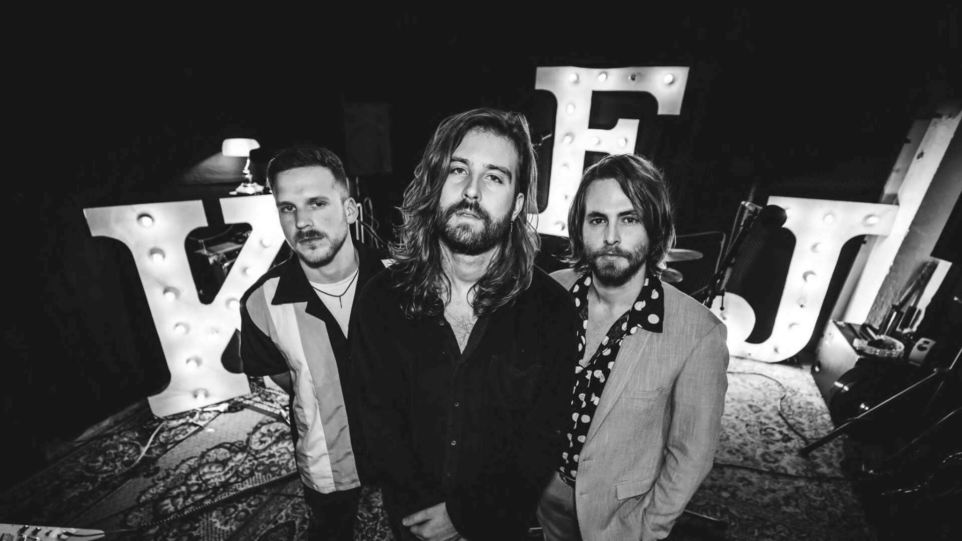 Die 3 Mitglieder der Band posieren in einem Proberaum, vor den Buchstaben K F und J. Das Bild ist in schwarzweiß