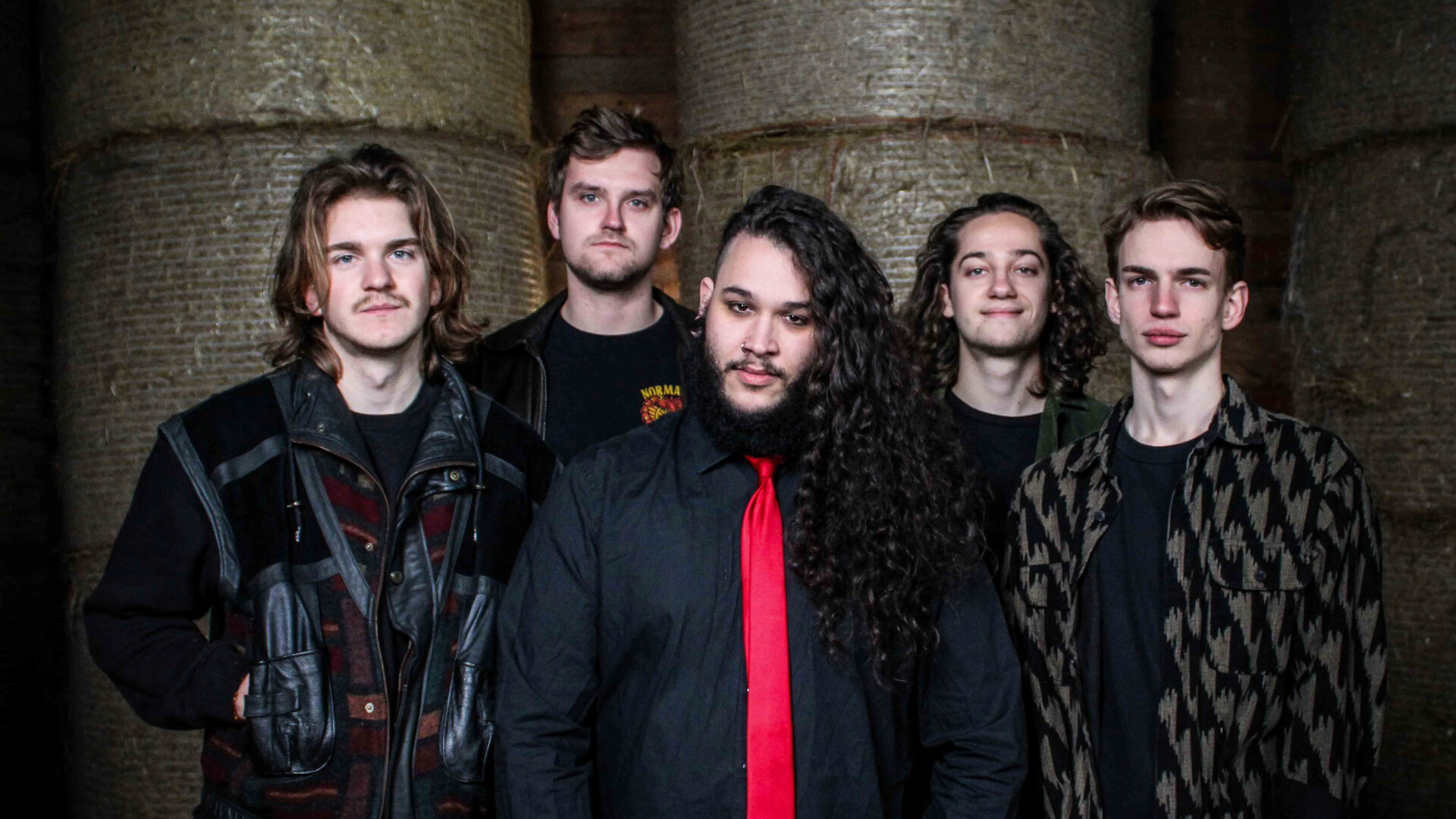 Die Band One Last Glance posieren in schwarzer beziehungsweise dunkel gehaltener Kleidung vor kaum Erkennbaren Heuballen hinter Ihenen. Der Sänger trägt eine rote Krawatte