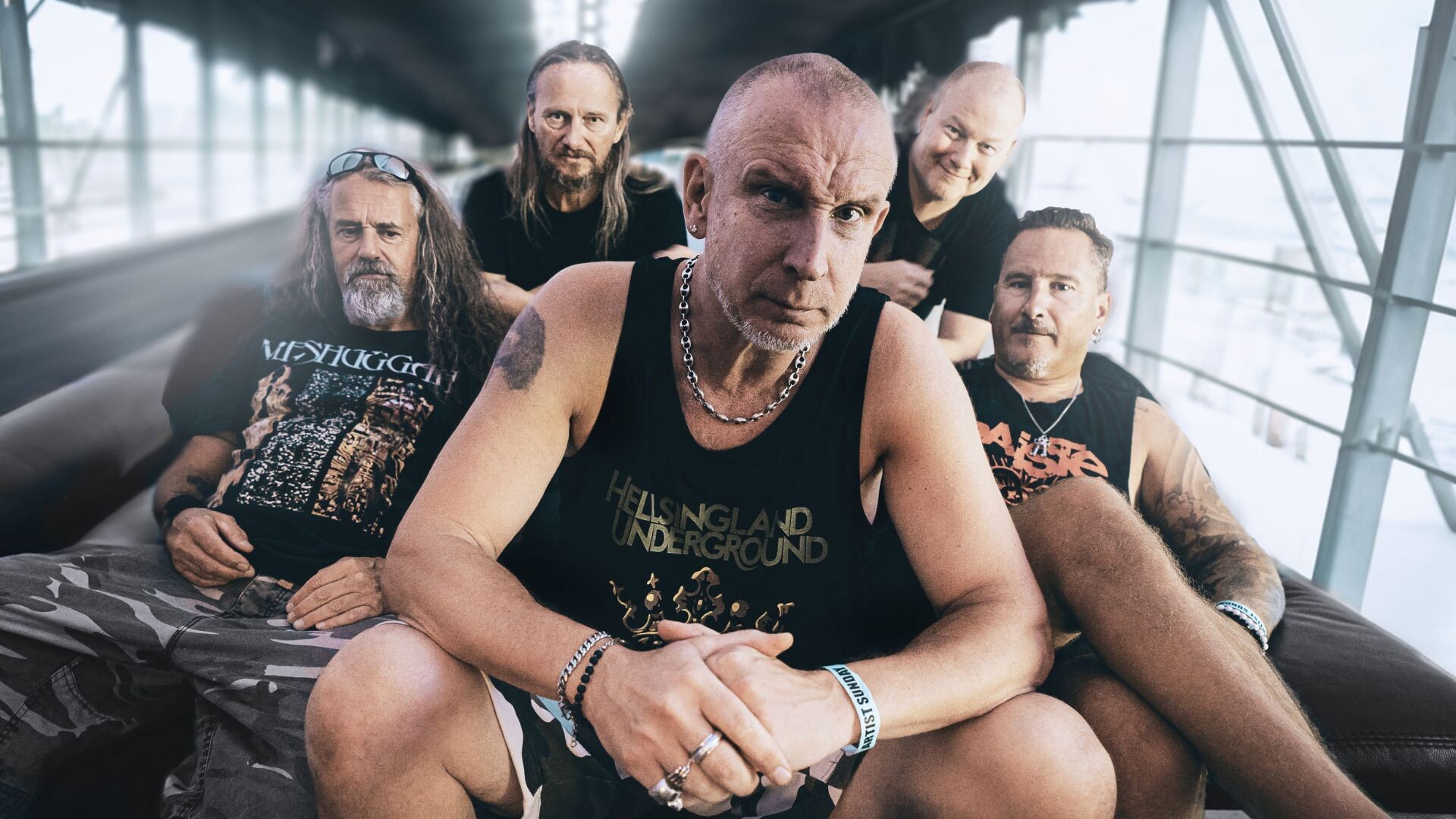Die Bandmitglieder von Clawfinger sitzen auf einer Couch in einem Langen Gang mit Fenstern auf den Seiten
