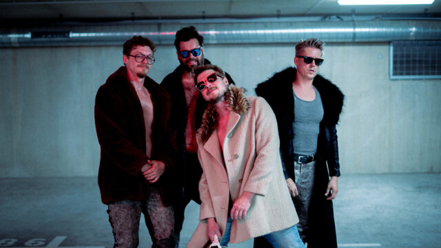 Die Band Old Mrs. Bates posiert in einem einer Tiefgarage ähnlich wirkendem Raum. Sie tragen offene Mäntel mit Shirts darunter.