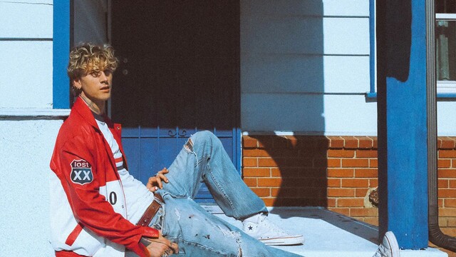 MGK sitzt vor einem Hauseingang, er trägt Jeans und eine Rote College Jacke