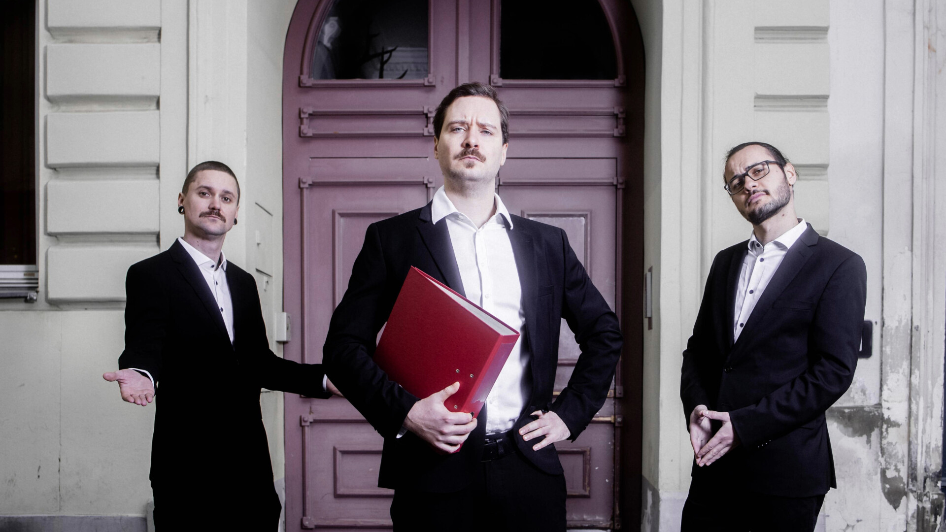 Die Band Steht in Anzügen vor einem Altbau mit rotem Tor